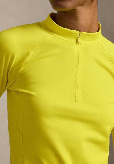 Κίτρινο τοπ με μακριά μανίκια, ψηλό γιακά και ημίψηλο φερμουάρ. Διαθέτει λείο, εφαρμοστό σχέδιο με διακριτικές λεπτομέρειες της μάρκας στο ύφασμα.