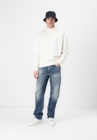 Sweat à capuche blanc, jean bleu déchiré et baskets blanches. Le modèle porte un chapeau seau noir. Tenue décontractée sur un fond uni.