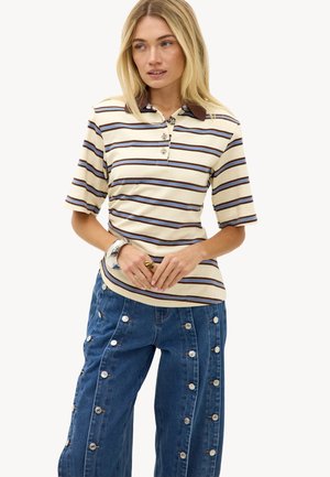 Vrouw die een beige, bruin en blauw gestreept poloshirt met korte mouwen draagt en wijde blauwe jeans met verticale knopen.