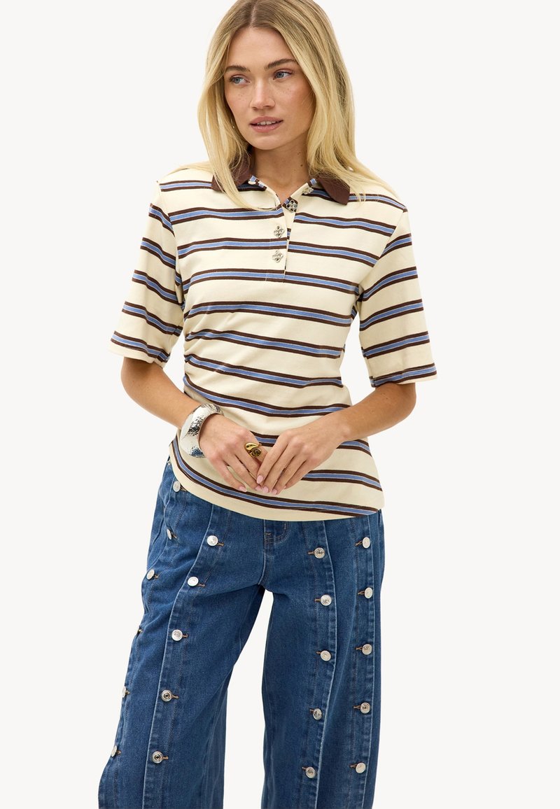 Femme portant un polo à manches courtes rayé beige, marron et bleu, avec un jean bleu à jambes larges orné de boutons verticaux.