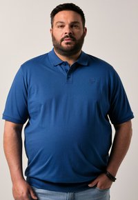 Modrá polo košile vyrobená z hladkého materiálu, s klasickým límečkem, třemi knoflíky na zapínání a decentním logem na hrudi.