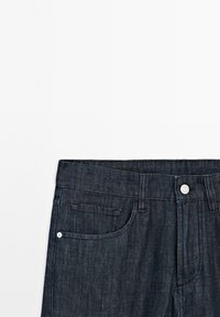 Jeans in denim scuro con vita alta, una chiusura a bottone singolo e una piccola tasca frontale visibile su uno sfondo bianco semplice.