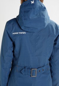 Blaue wasserdichte Jacke mit Kapuze, tailliertem Schnitt mit silberner Gürtelschnalle und dem Logo „KARI TRAA“ auf der Rückseite. Glatte Textur.