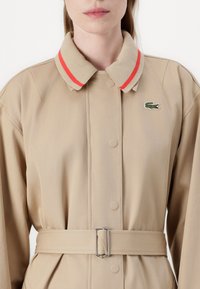 Cappotto beige con colletto beige chiaro, caratterizzato da strisce rosse, una finitura testurizzata e una cintura con fibbia argentata; include un logo ricamato verde.