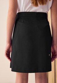 Femme portant une jupe noire au genou et un chemisier blanc, debout les mains reposant naturellement le long du corps, sur un fond neutre.