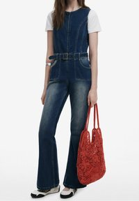 Kvinde iført mørk denim jumpsuit med bælte og hvid T-shirt, holder en rød blomsterhæklet taske, stående mod en hvid baggrund.