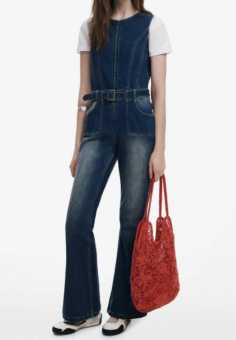 Kvinde iført mørk denim jumpsuit med bælte og hvid T-shirt, holder en rød blomsterhæklet taske, stående mod en hvid baggrund.