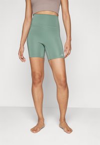 Shorts de sport taille haute, verts, avec une texture lisse et des coutures contrastées. Logo New Balance en gris visible sur la jambe gauche.