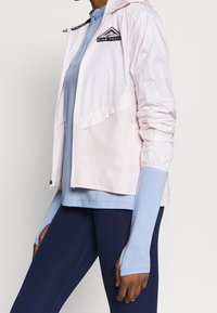 Leichte, rosa-weiße Windjacke mit Kapuze, silbernen Akzenten und einem dreieckigen Logo. Trägt man über einem blauen, geschichteten Oberteil und dunklen Leggings.