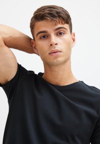 T-shirt di cotone nero con scollo rotondo, design semplice e texture liscia. Modello posa con una mano dietro la testa, sfondo neutro.