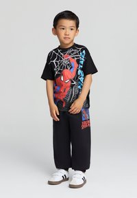 Maglietta nera con una grafica di Spider-Man rossa e blu, abbinata a pantaloni neri larghi con un logo stampato. Sneakers bianche con accenti scuri.