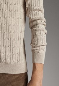 Beige-värinen kaapelikudottu neule, joka esittelee tekstuurillisen kuviion, resorit hihansuissa ja istuvan mallin, yhdistettynä ruskeisiin pörrökangas-housuihin.