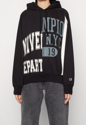 Osoba ubrana w czarną bluzę z kapturem z dużym, mieszanym białym i niebieskim napisem, zawierającym słowa "NYVER", "MPIO", "N.Y.C" oraz "19", w połączeniu z szarymi dżinsami.