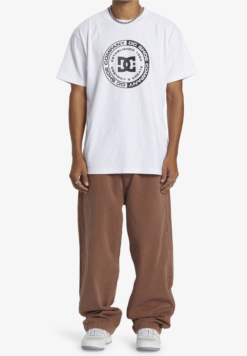 Vit bomullst-shirt med en cirkulär svart grafik med text, kombinerad med bruna byxor med ledig passform och grå sneakers.