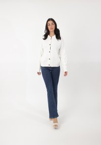 Cárdigan blanco de punto con botones marrones, con cuello redondo y puños acanalados, combinado con jeans acampanados azul oscuro y sandalias blancas.