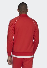 Red athletic jacket with white accents, long sleeves, and a stand-up collar. Features a smooth texture and ribbed cuffs and hem.
