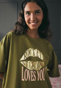 Tricou oversized de culoare verde măslin, cu mâneci scurte. Prezintă un text grafic metalic în roz și auriu: "ROCKETT ST GEORGE TE IUBEȘTE" cu contur de buze.