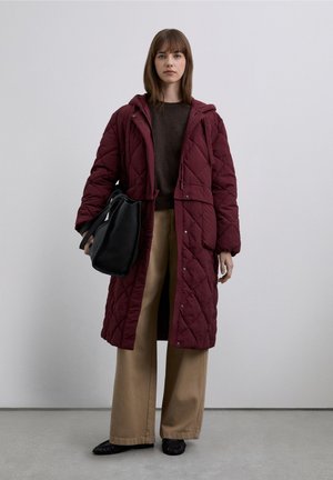 Abrigo largo acolchado color burdeos con capucha, combinado con un suéter marrón y pantalones beige de pierna ancha. La modelo sostiene un bolso negro.