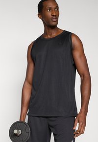 Μαύρο αθλητικό τοπ χωρίς μανίκια, κατασκευασμένο από διαπνέον ύφασμα διχτυού, με στρογγυλή λαιμόκοψη και λεπτομέρεια λογότυπου. Το μοντέλο κρατάει ένα μαύρο βάρος.