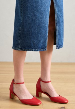 Punased nahast kontsad, ümmarguse varbaga, pahkluu rihmaga ja plokknõgaga. Sile tekstuur kuldse tooni aktsentiga kontsal.