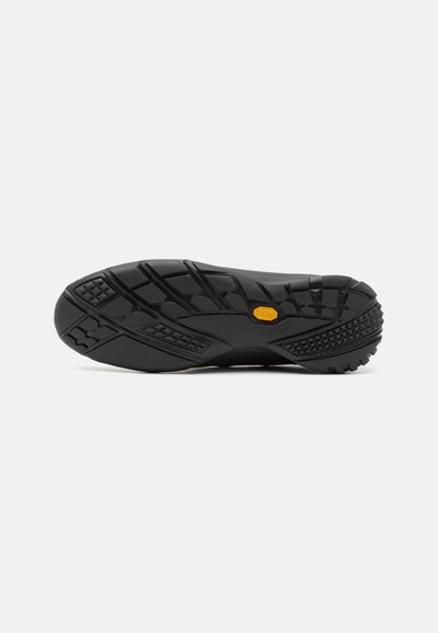 Semelle de chaussure en caoutchouc noir avec des motifs de texture, comportant un logo de marque jaune bien visible et plusieurs zones de grip pour l'adhérence.