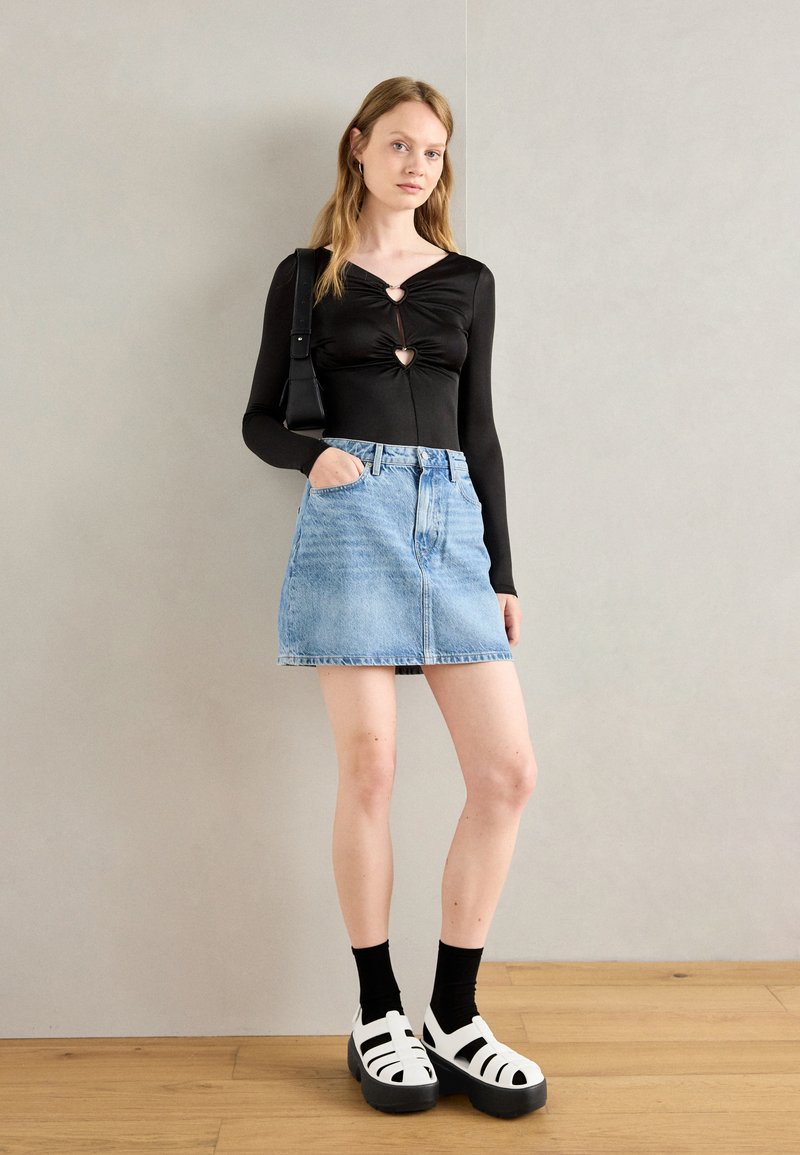 Schwarzes Langarmshirt mit vorderem Knoten-Detail, hellblauen Jeansrock, schwarzen Socken und weißen Plateausandalen mit schwarzen Sohlen.