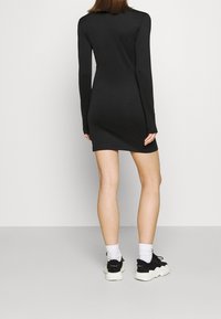 Person med långärmad svart miniklänning, vita strumpor och svartvita sneakers, står mot en neutral bakgrund med ryggen vänd mot kameran.