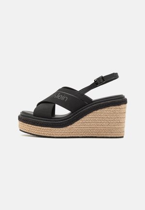 Sort kile-sandal med flettet halmplatform, krydsede elastiske stropper, justerbar bagrem og subtil Calvin Klein-logo på øverste rem.
