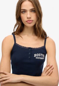 Marineblauwe geribde tanktop met kantafwerking, voorzien van knoopsluiting en de tekst "BOSTON SUPERDRY" in het wit. Verstelbare spaghettibandjes.