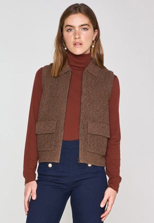 Brown knitted vest with ribbed texture, has two front pockets, worn over a brown turtleneck, paired with dark blue pants.