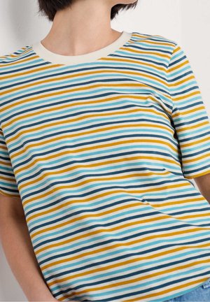Person iført en flerfarvet T-shirt med hvide, gule, blågrønne og mørkeblå vandrette striber mod en ensfarvet baggrund.