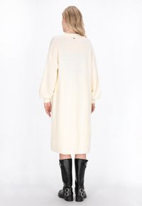 Weißes, oversized gestricktes Kleid mit langen Ärmeln, gerippter Textur und seitlichen Schlitzen, kombiniert mit schwarzen kniehohen Stiefeln mit Schnallen.