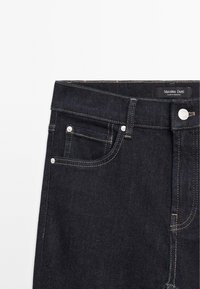 Tmavé džíny s klasickým střihem, které mají malou přední kapsu, stříbrné zapínání na knoflík a kontrastní detaily žlutého prošívání.