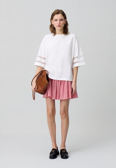 T-shirt bianca oversize con dettagli a righe, gonna plissettata a scacchi rossi e bianchi, borsa marrone e mocassini neri.