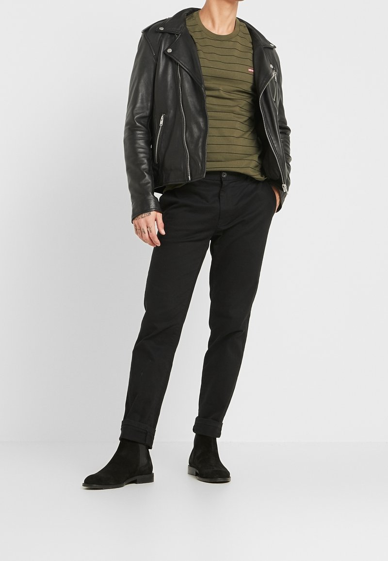 Homme portant une veste en cuir noire, un t-shirt à rayures vert olive, un pantalon noir et des bottes noires, debout devant un fond uni.