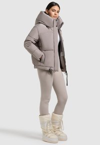 Donsjack in gedempt grijs met een hoge kraag, rits aan de voorkant en zakken. Gecombineerd met zachte beige leggings en witte sneeuwboots.