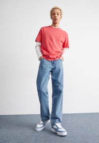 Koraalkleurig katoenen t-shirt over een wit longsleeve shirt, gecombineerd met lichtblauwe denim jeans en wit-blauwe sneakers. Effen achtergrond.