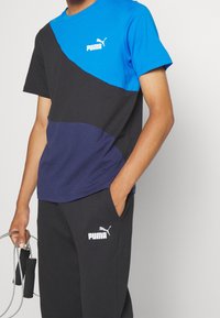 Pánský sportovní outfit se skládá z trička v barevných blokacích modré, černé a námořnické barvy a černých kalhot s logem, vyrobeného z měkké látky.