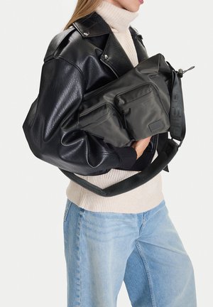 Osoba v černé kožené bundě, béžovém roláku a světle modrých džínách, držící velkou černou tašku přes rameno s předními kapsami a nastavitelným páskem.