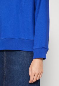 Sudadera azul con puños y dobladillo acanalados, que presenta una textura suave. Mano visible, sosteniendo una tela de mezclilla.