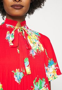 Röd blommig klänning med hög krage, har ett knytband, korta ärmar och strukturerad veckning. Ljusa, flerfärgade blommönster som accentuerar.