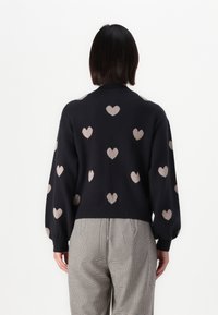 Maglione blu scuro con motivi a cuori rosa pallido, caratterizzato da un collo rotondo e maniche a palloncino. Indossato con pantaloni a quadri.