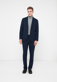 Jovem de pé, vestindo um fato azul-marinho com um suéter de pescoço alto cinza e mocassins pretos, contra um fundo branco simples.