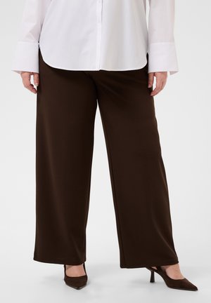 Personne portant un pantalon large marron foncé, une chemise blanche à manches longues boutonnée et des talons pointus marron foncé, debout sur un sol blanc.