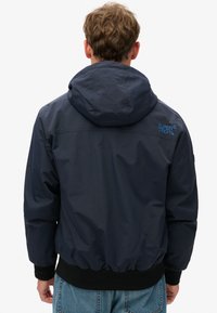 Marineblå letvægtsjakke med hætte, elastiske manchetter og talje, har et logo i lyseblå på den øverste ryg, glat tekstur.