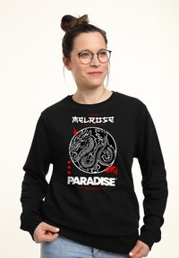 Černá mikina s bílou grafikou draka a červeným textem; kulatý design s propracovanými detaily; žebrované manžety a lem.