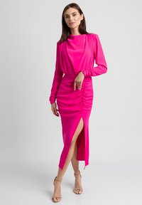 Fuksjowa sukienka z długim rękawem, z drapowanym dekoltem, marszczonymi bokami i rozcięciem na wysokości uda. Gładki materiał, obcisły krój i minimalne akcenty.