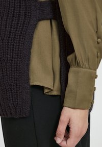 Strikket mørkelilla sweater lagdelt over en olivengrøn bluse med samlede ærmer og knapdetaljer, delvist synlig ved håndleddet.