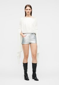 Γυναίκα με καστανά ίσια μαλλιά που φοράει λευκό μπλουζάκι χωρίς ώμους, ασημένια σορτς και μαύρες μπότες μέχρι το γόνατο, στέκεται μπροστά σε λευκό φόντο.