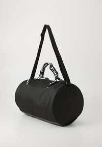 Sac de sport cylindrique noir avec fermeture éclair, comportant des anses tissées noires et blanches avec le logo "PUMA". Fabriqué en tissu texturé.